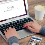 Hands typing on MacBook Air with Google search open, coffee nearby.