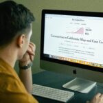 A man reading a coronavirus case count article on a computer screen indoors.